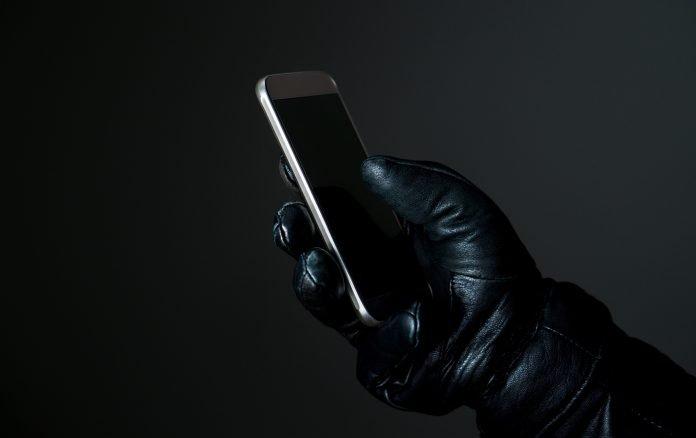 Mobile and cyber security thread concept. Criminal holding smartphone with black leather gloves in darkness. Hacker touching smart phone screen. Phone scam