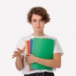 Unhappy young student in a white shirt and eyeglasses holding colorful folders showing so-so gesture